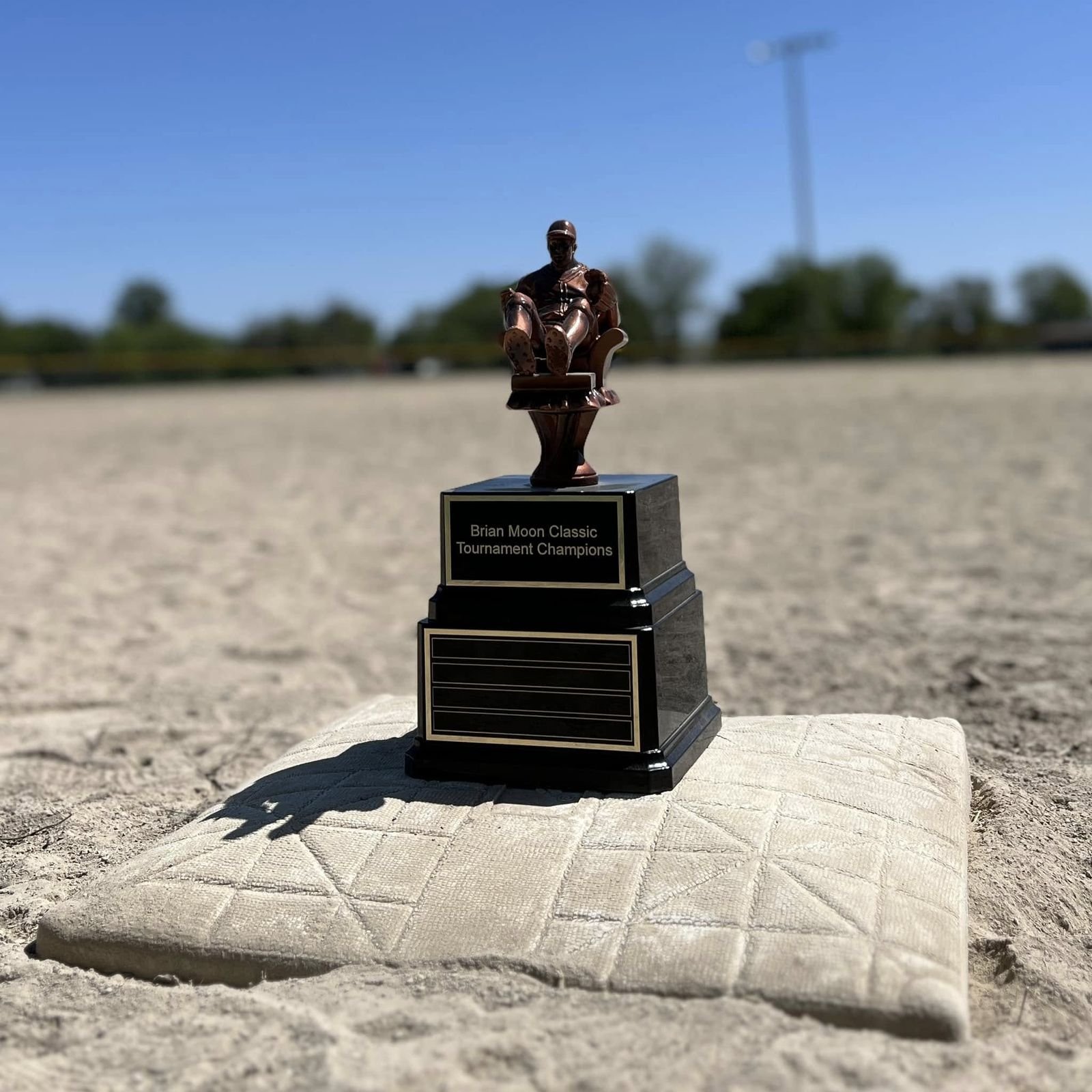 The Brian Moon Classic traveling trophy, a bronze figure seated in a recliner, standing on a base bag on the infield dirt. The plate reads Brian Moon Classic Tournament Champions.